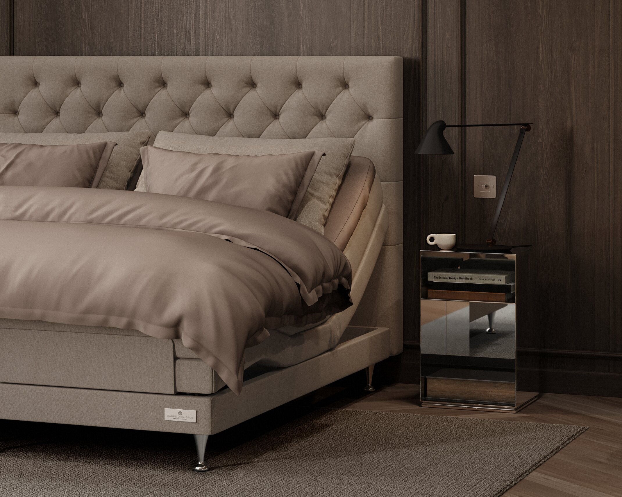 A modern bedroom scene featuring a bed with a tufted beige upholstered headboard, matching beige bedding, a dark wood-paneled wall, a small mirrored nightstand with a black lamp, and a gray rug.