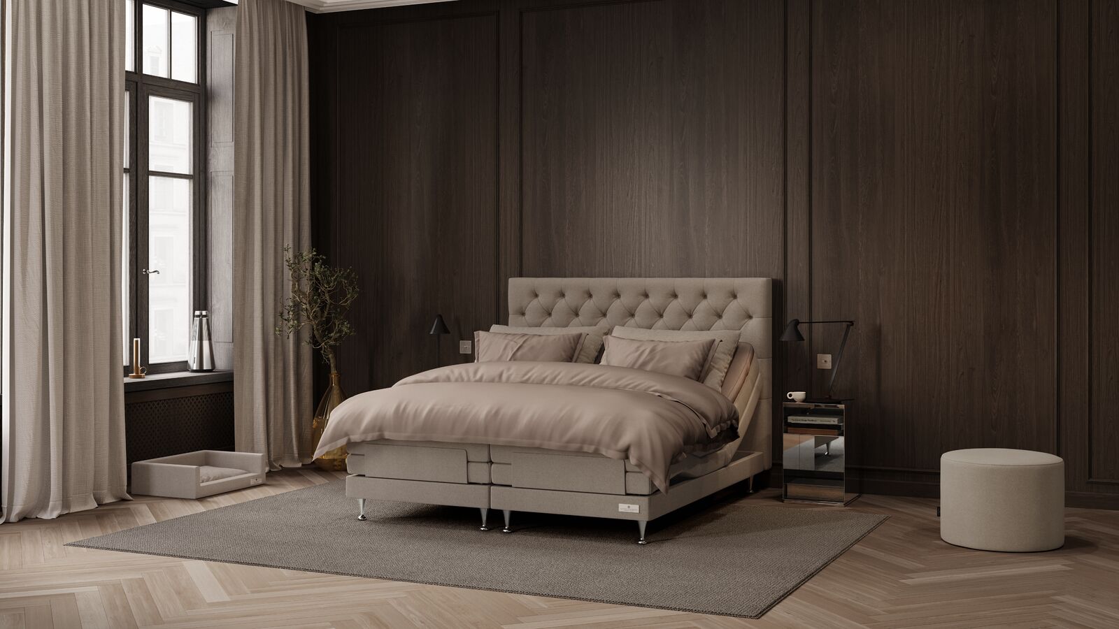 A modern bedroom featuring a tufted beige platform bed with matching bedding, set against a dark wood-paneled wall with a window, curtains, side tables, and a round ottoman.