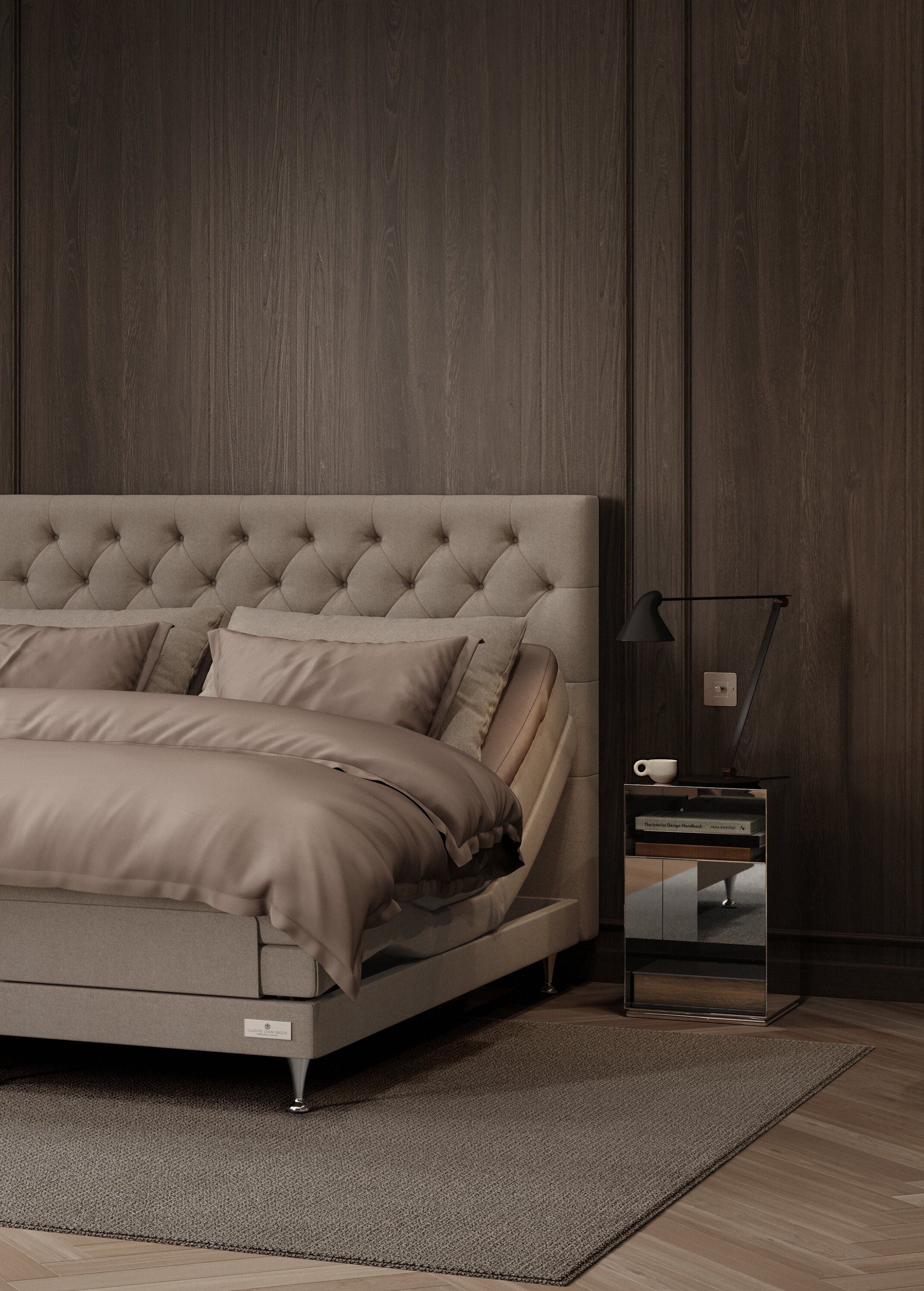 A modern bedroom scene featuring a bed with a tufted beige upholstered headboard, matching beige bedding, a dark wood-paneled wall, a small mirrored nightstand with a black lamp, and a gray rug.