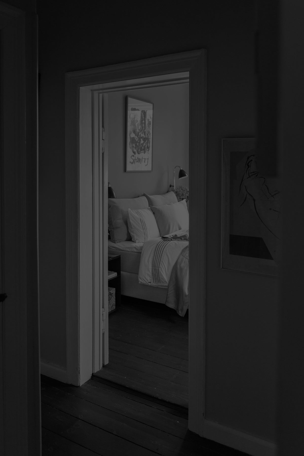 A dark, moody interior photograph looking through an open doorway into a bedroom. The room contains a bed with white bedding, a framed print on the wall, and a lamp.